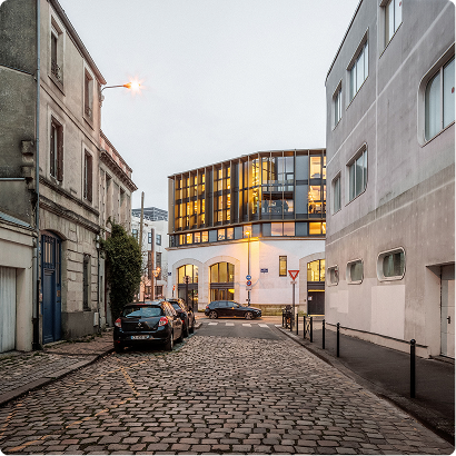 Vue urbaine d’un bâtiment contemporain éclairé, situé au bout d’une rue pavée - locaux BPM Architectes